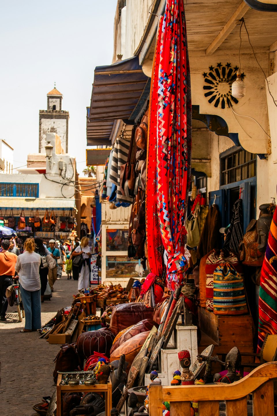 Shoppers walk past textile stalls and leather goods on a cobblestone street.